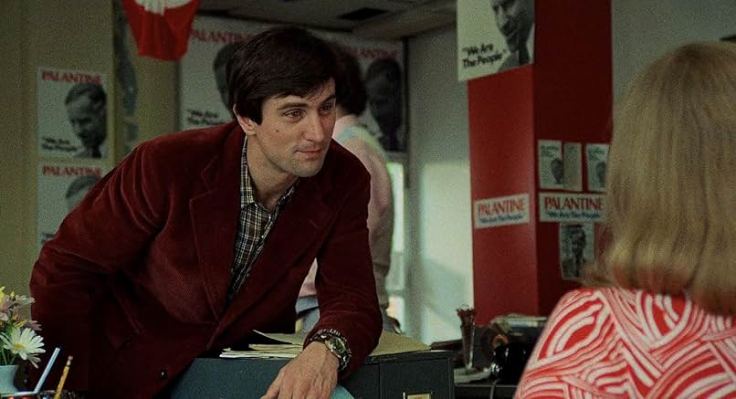 Robert De Niro and Cybill Shepard, Travis leans over Betsy's desk casually flirting and wearing a red blazer.