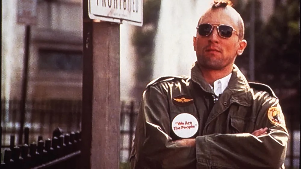 Travis Bickle is listening to Palpatine's speech, wearing a green field jacket, a campaign button that says "We Are The People," aviator sunglasses, and his head is shaven in the style of a mohawk.